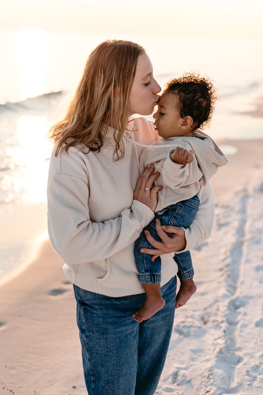 Mother kissing her son during golden hour on the beach with perfect white sand and golden glow.