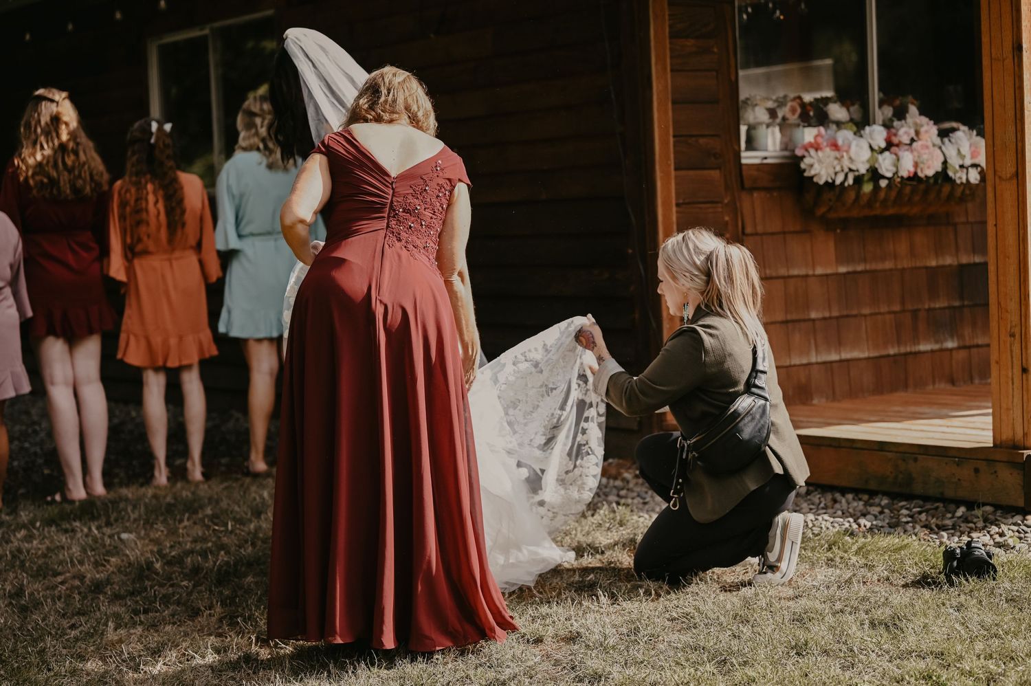 Two women in formal dresses examine delicate white lace fabric outside a rustic wooden building.