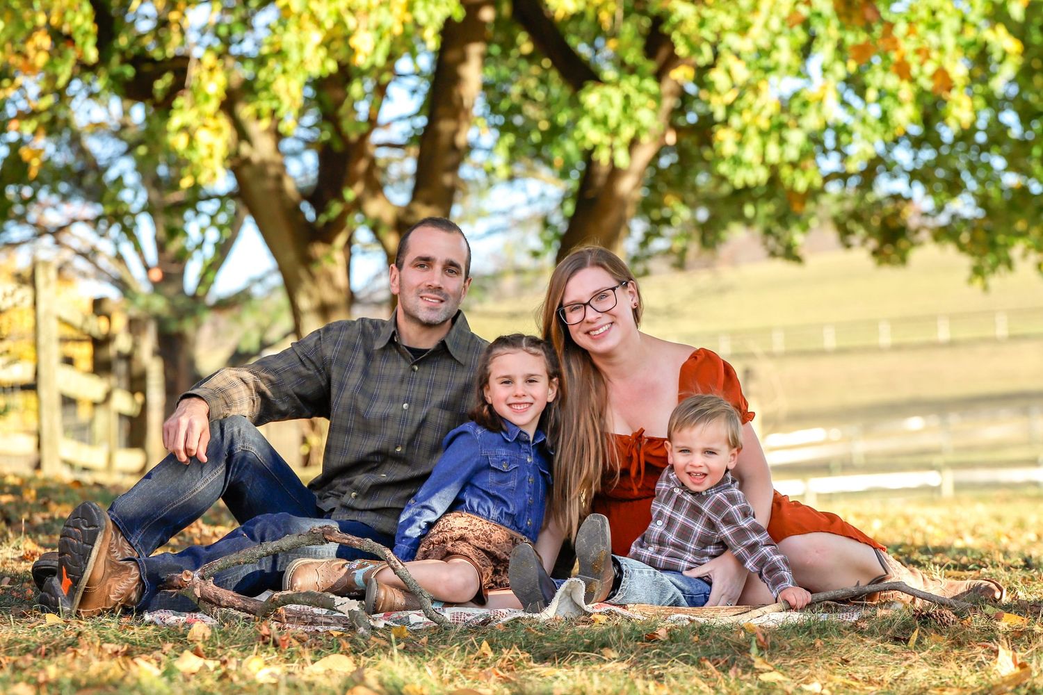 Family sits together on blanket under autumn tree for outdoor portrait session at golden hour.