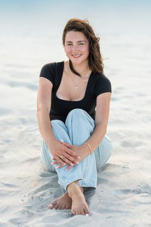 Young woman in black top and jeans sitting barefoot on white sand beach, smiling warmly at camera.