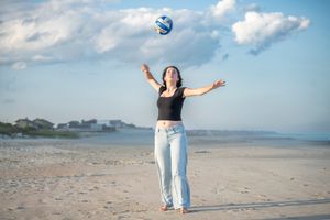 Woman in black top and jeans tossing a volleyball on a sandy beach under a partly cloudy sky.