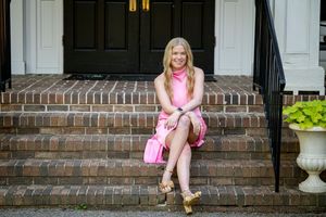 A person in a pink dress sits on brick steps leading to a white-columned entrance, with a matching pink purse nearby.