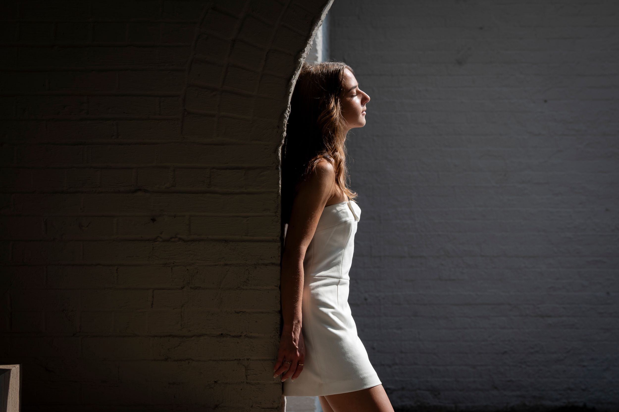 Woman in white mini dress posing against dark brick wall in dramatic side lighting.