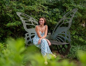 A person in a floral dress sits on a butterfly-shaped garden bench surrounded by lush greenery.