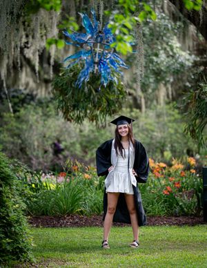 Graduate in cap and gown poses in a garden with Spanish moss and vibrant blue flowers overhead.