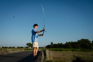 A young boy in a blue shirt casts a fishing rod while standing on a dock at sunset.