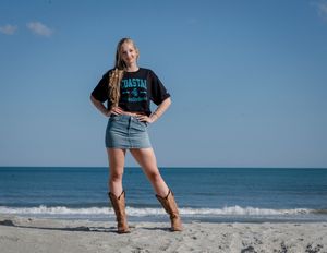 Person wearing blue t-shirt and denim skirt poses confidently on sunny beach with ocean waves in background.