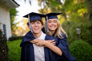 Two college graduates in navy blue caps and gowns smile together in an outdoor garden setting.