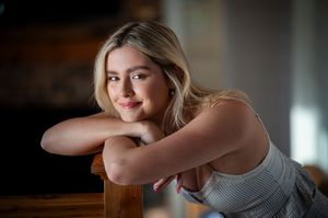 Portrait of a young woman in a white tank top leaning on a wooden surface in soft indoor lighting.