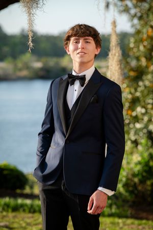 A person in a formal black tuxedo and bow tie poses outdoors by a lake with lush foliage in the background.