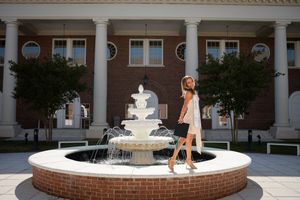 Young girl posing by a white tiered fountain in front of a classic brick building with white columns.