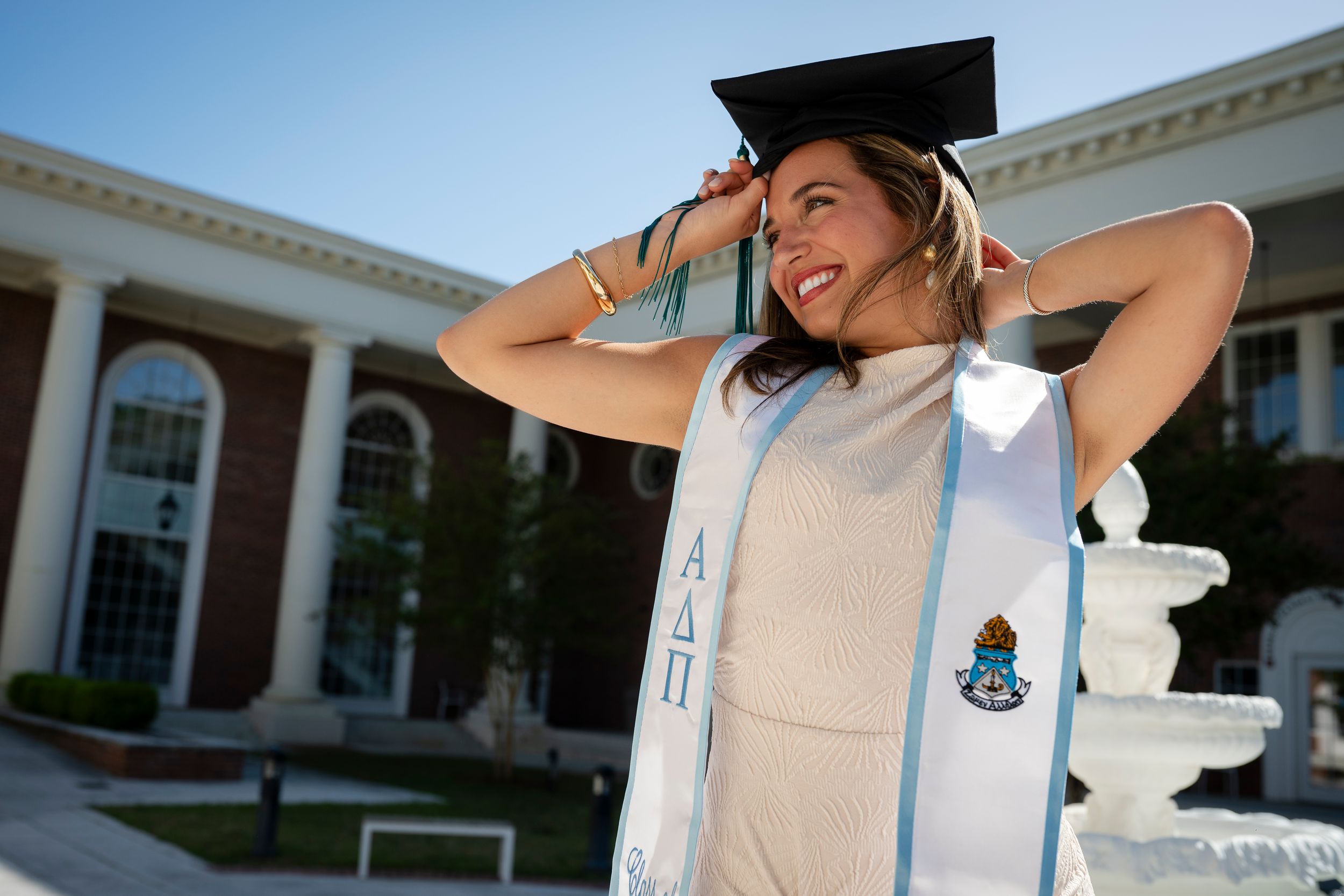 Smiling college graduate in cap and stole poses outdoors on campus on a sunny day.
