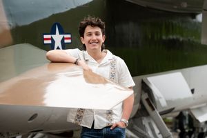 A smiling person in a white uniform shirt stands next to a WWII era military aircraft.