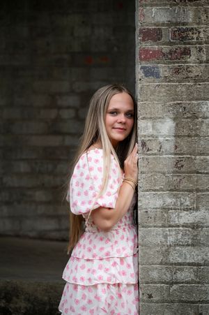 A person in a pink heart-patterned dress leans against a gray brick wall while looking at the camera.