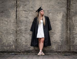 Graduate in cap and gown leans against concrete wall wearing white dress and casual sneakers.