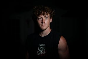 A studio portrait against black background shows someone in a sleeveless dark t-shirt with curly hair.