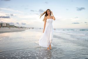 Person in white dress walks barefoot along misty beach shoreline during sunset.