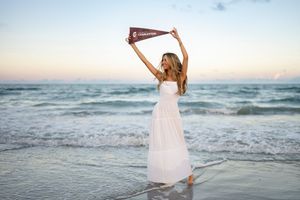 A white dress-clad figure stands in ocean waves at sunset holding up a pink pennant flag against a pastel sky.