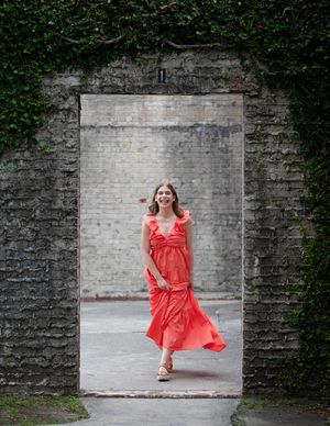 A figure in a flowing coral red dress moves dynamically against a stone wall backdrop creating an artistic motion effect.