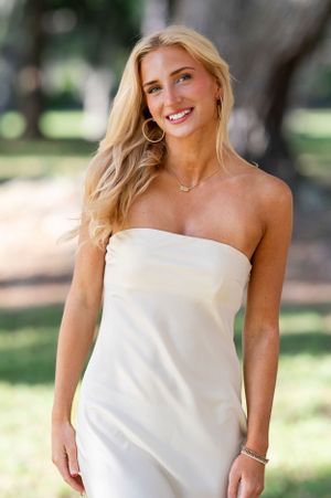 A person in a white strapless dress poses outdoors with natural lighting and a blurred background.