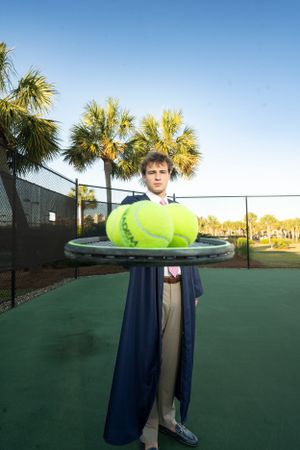 Person holding oversized tennis ball props on outdoor tennis court with palm trees.
