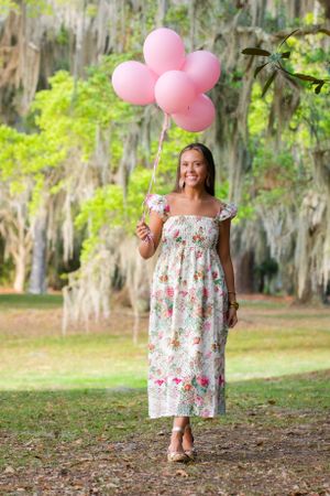 A floral dress-clad figure holds pink balloons in a scenic park with Spanish moss.