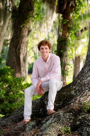 A person in a plaid button-down shirt and khakis sits casually on a mossy tree trunk with Spanish moss hanging overhead.