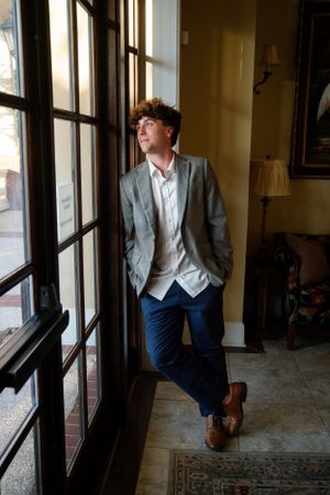 A person in business casual attire leans against a large window frame in a traditional room setting.