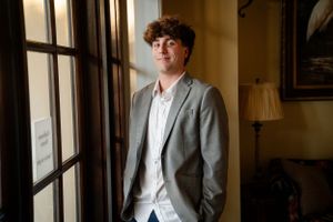 A person in a grey blazer and white shirt stands pensively by a window in an elegant interior.