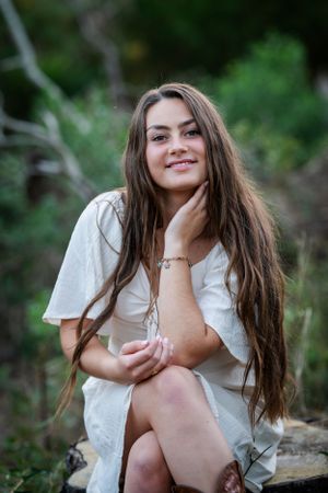 A long-haired figure in a white dress sits outdoors in a natural setting with greenery in the background.