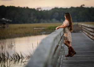 Woman in floral dress leans on railing of coastal boardwalk at sunset.