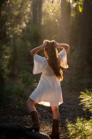 A person in a white dress and boots poses in a sun-drenched forest setting creating a dreamy backlit silhouette.
