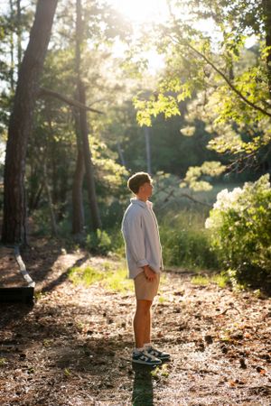 Person in light clothing stands in sunlit wooded area.