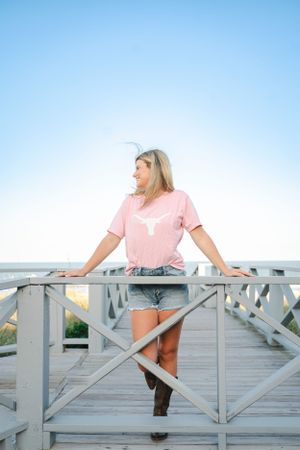 Standing on a wooden boardwalk in a pink shirt and denim shorts with ocean view.