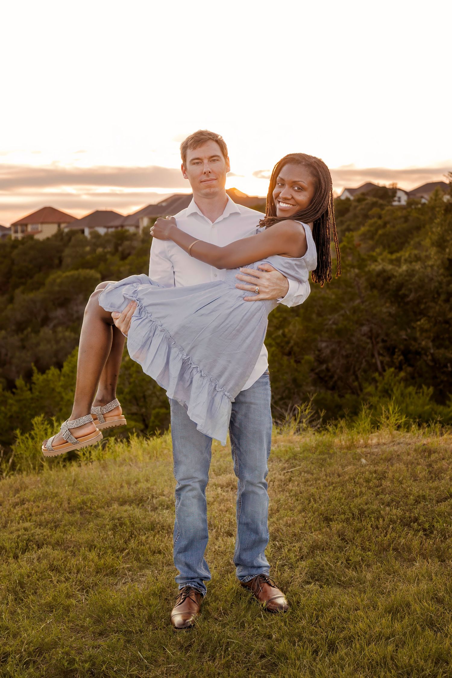 A couple embraces during sunset on a grassy hill, with the woman wearing a flowing white dress and being held aloft.