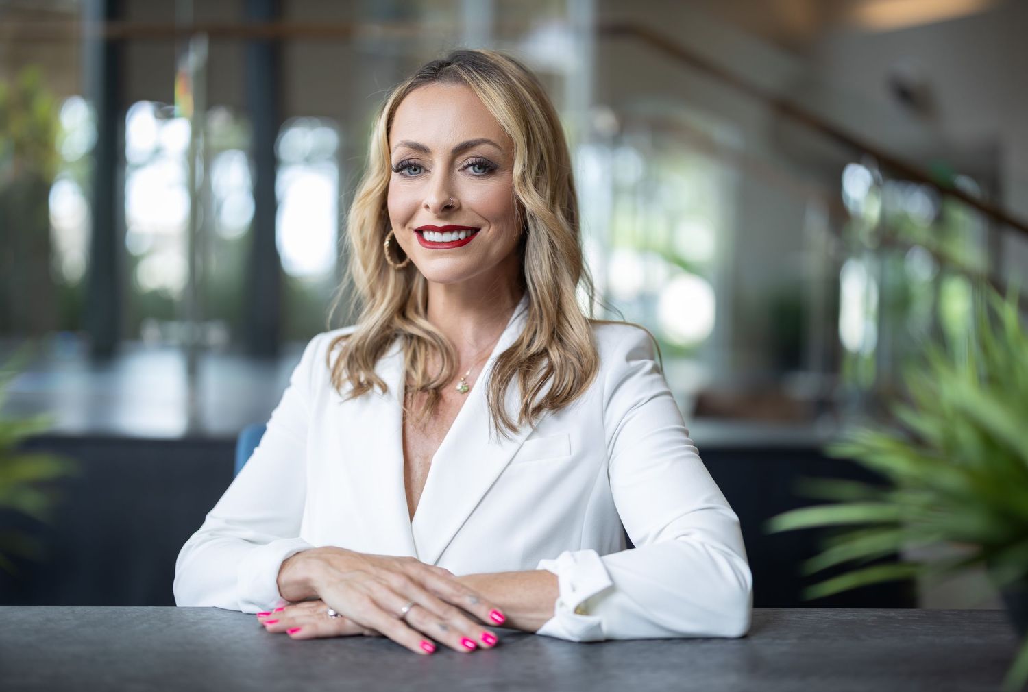 Professional portrait of someone in white blazer sitting at desk in modern office setting.