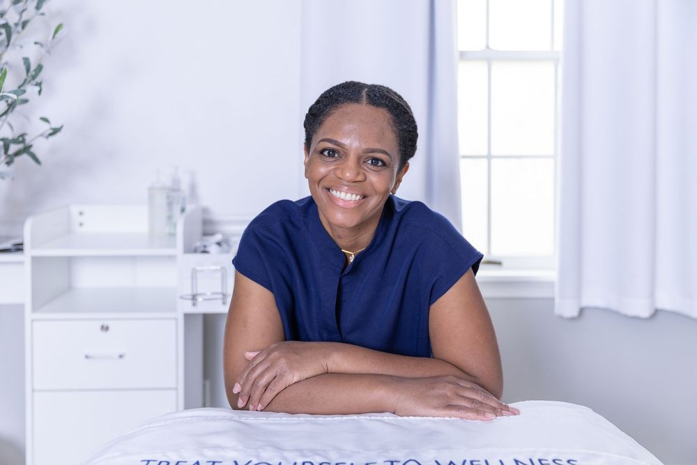 A person in a navy blue shirt sits at a desk in a bright office space, smiling brightly at the camera.