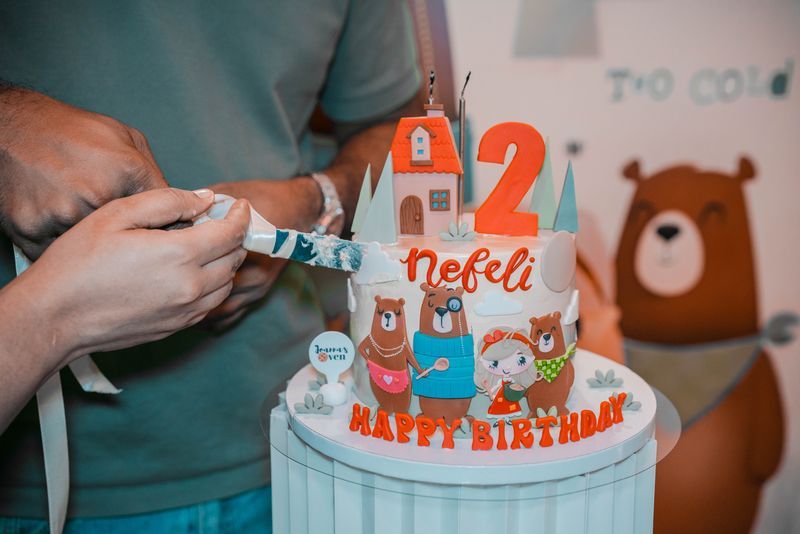 Hands lighting a birthday candle on a blue cake with a red number 2 topper and teddy bear decorations.