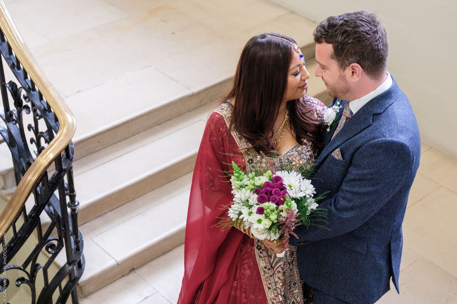 Couple in formal attire embracing on stairs as bride holds purple and white floral bouquet.