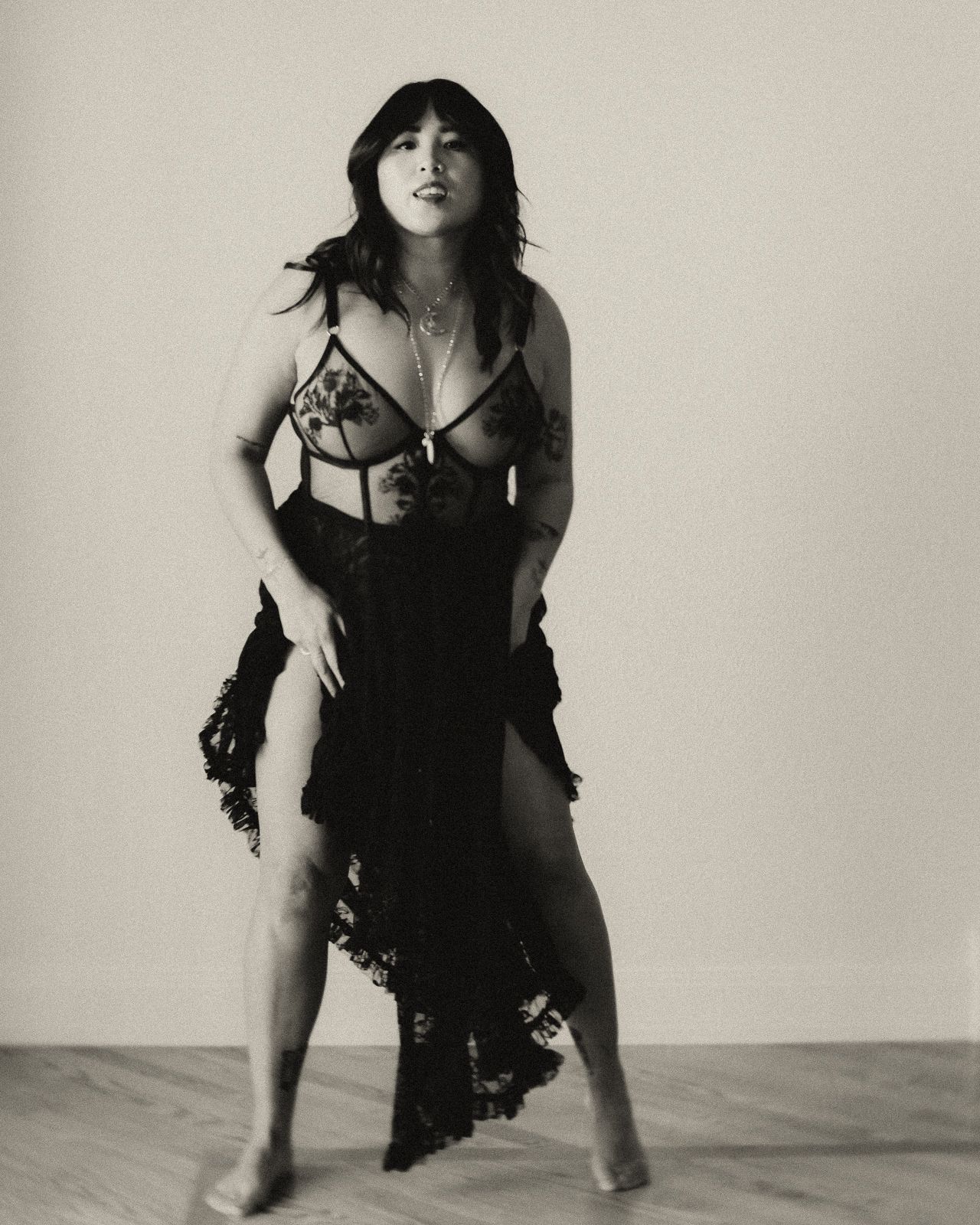 Black and white artistic portrait photograph of someone posing in a dark lace dress against a neutral background.