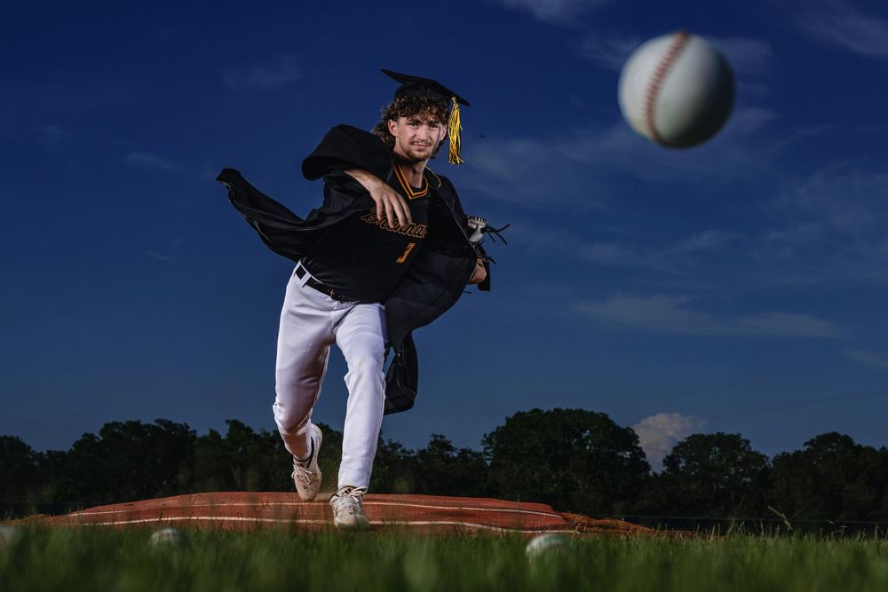 A baseball pitcher in mid-delivery on a mound at dusk with a baseball floating in the dark blue sky.