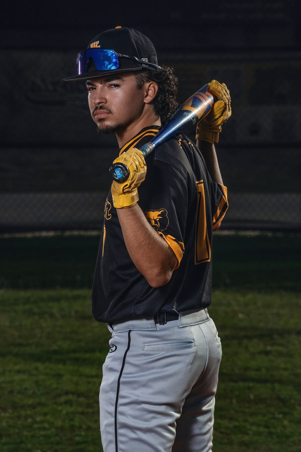 Baseball player in navy uniform and yellow batting gloves holds bat in ready stance against dark background.