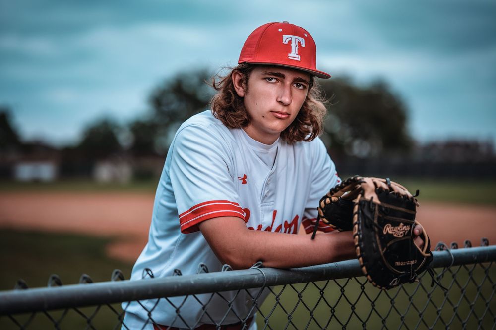 Baseball player in Texas Rangers uniform leans on chain link fence with glove at field during dusk practice session.