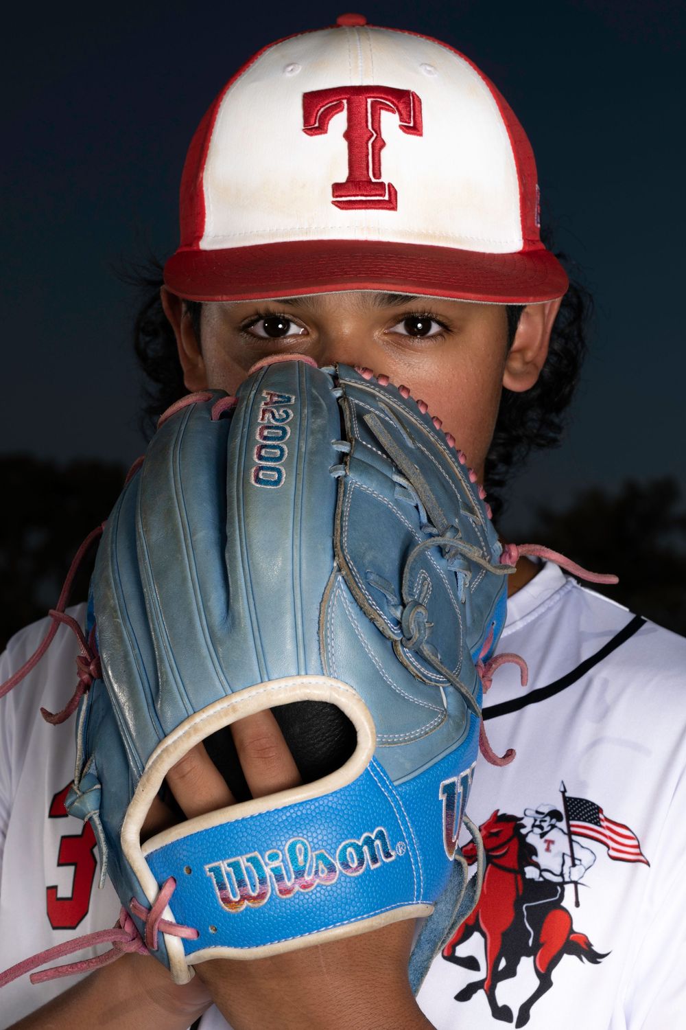 A baseball player wearing a Texas Rangers cap poses with a light blue Wilson glove covering part of their face.