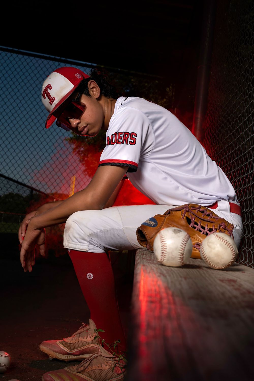 Baseball player in white uniform sits in dugout with glove and baseballs in dramatic red lighting.