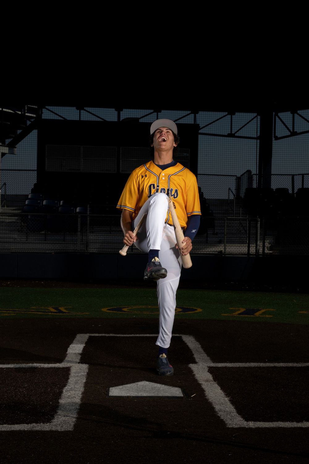 Baseball player in yellow jersey and white pants performing high leg kick pitching motion on baseball field at dusk.