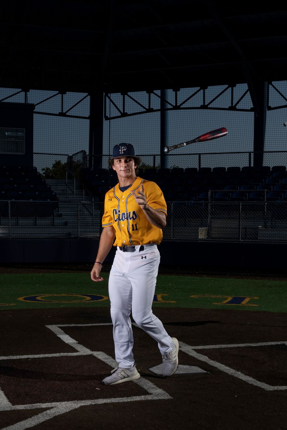 Baseball player in gold jersey and white pants poses with bat on home plate in dramatic stadium lighting.