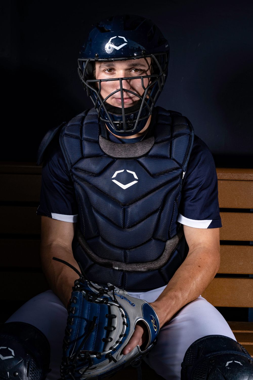 Baseball catcher wearing navy protective gear and helmet sits on wooden bench in dramatic lighting.