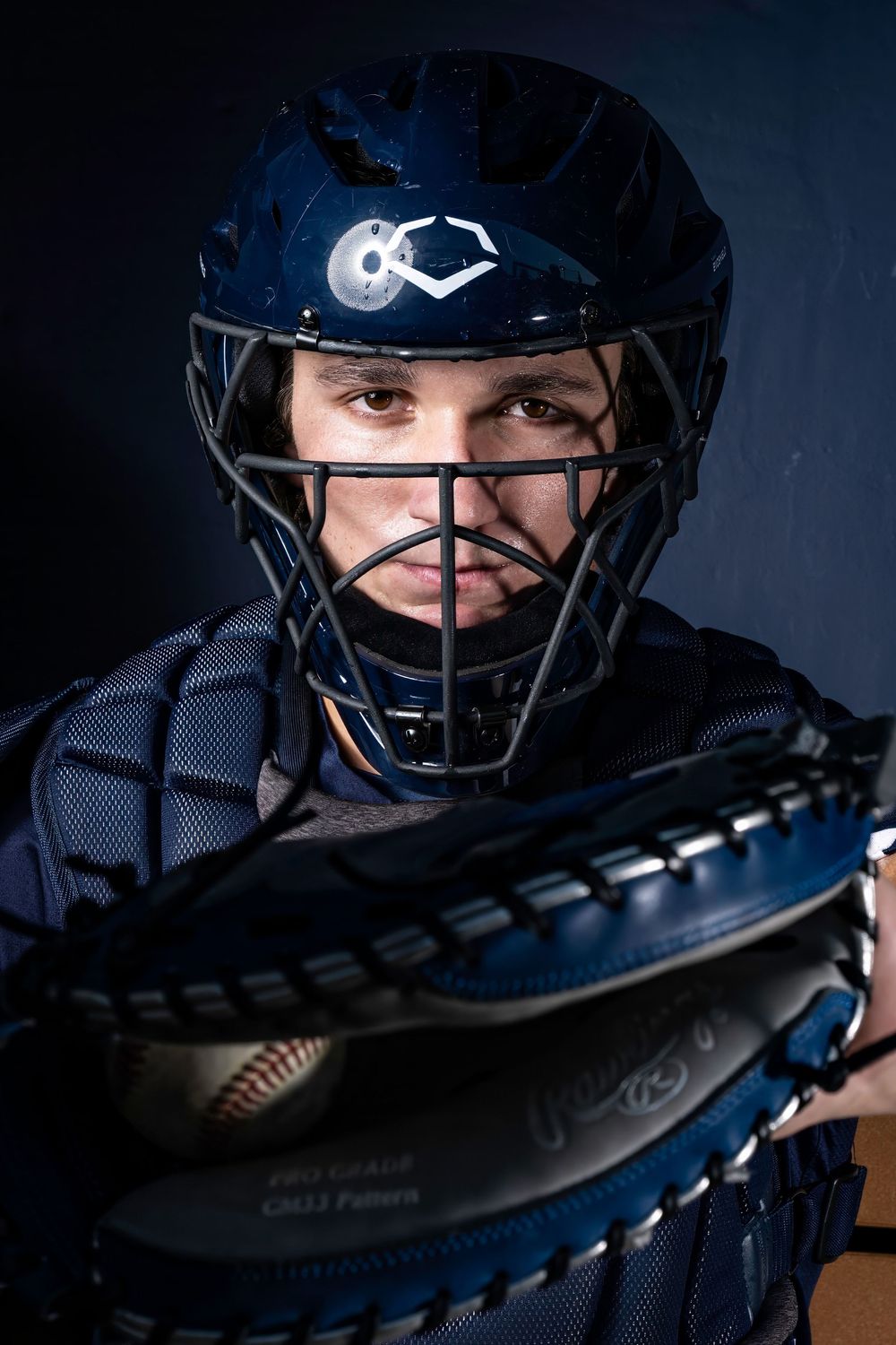 Baseball catcher in navy blue gear and helmet holding a mitt in dramatic lighting against dark background.