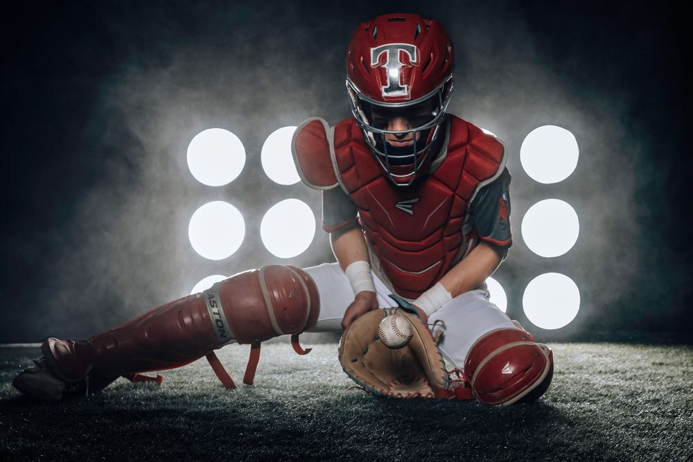 Baseball catcher in red gear crouches in position on field at night with stadium lights glowing in background.